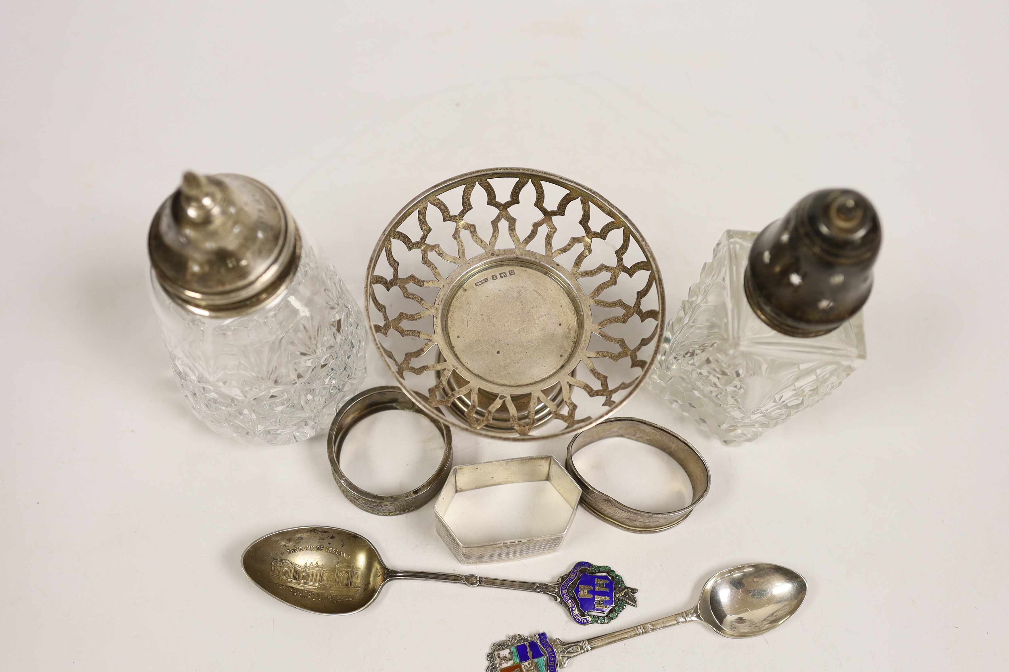 A group of small silver including two mounted glass sugar sifters, three assorted napkin rings, two commemorative spoons and a small pedestal nut dish (lacking insert).